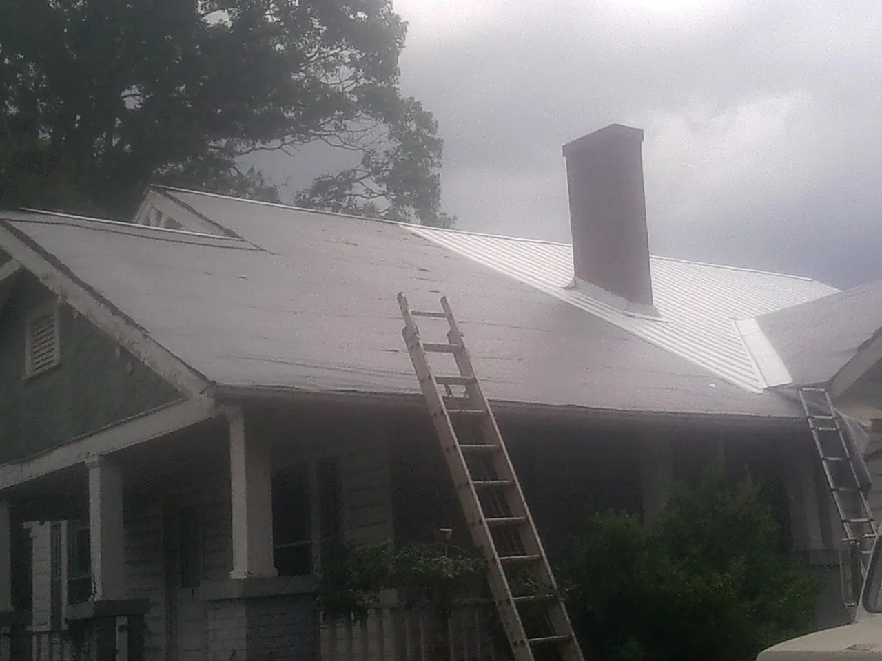 Ladders against a metal roof on an older home with a brick chimney in Manton